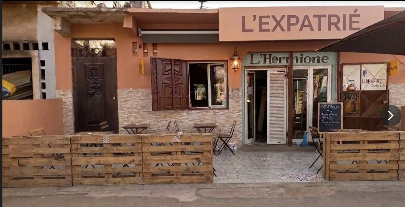 La façade de L'Expatrié, quartier Bâcha à Moroni : mur abricot, terrasse en bois et tables sur le trottoir.